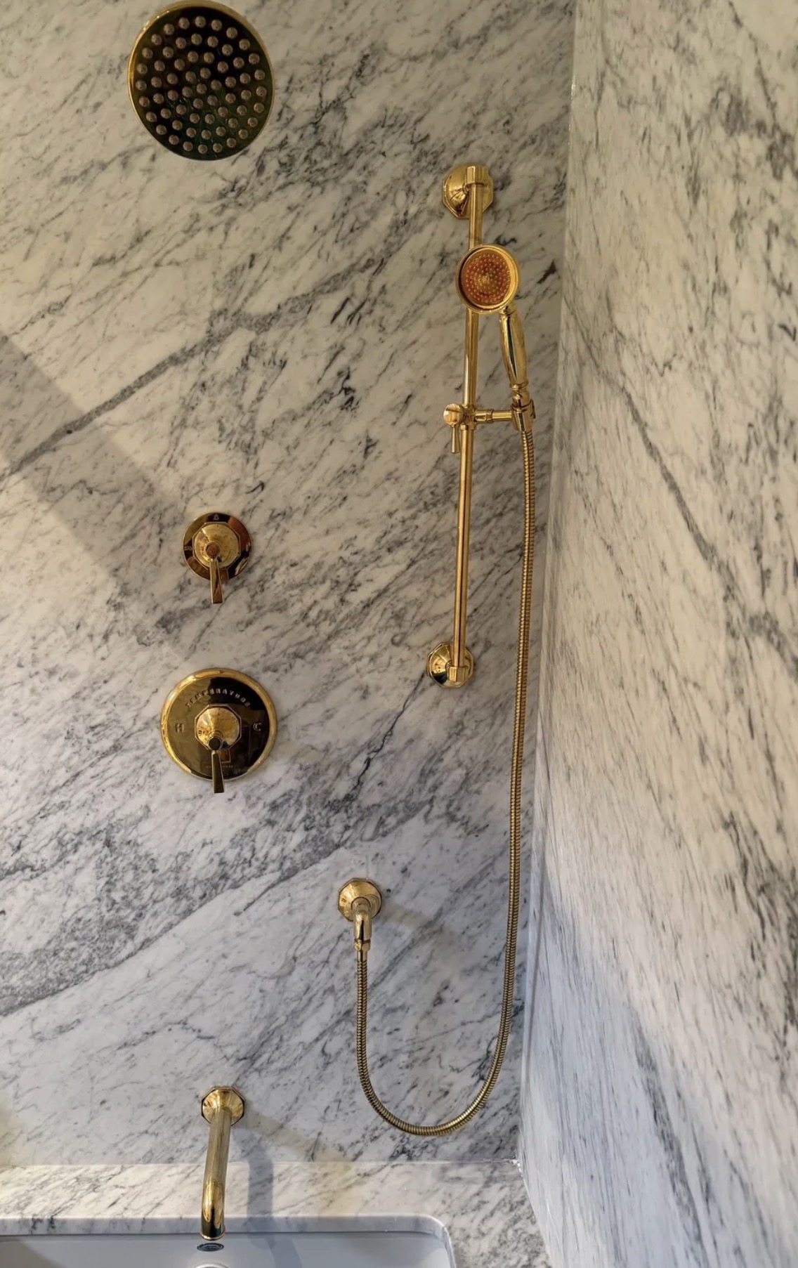 Polished brass rainfall shower head, hand shower, and tub filler installed on Carrara marble walls
